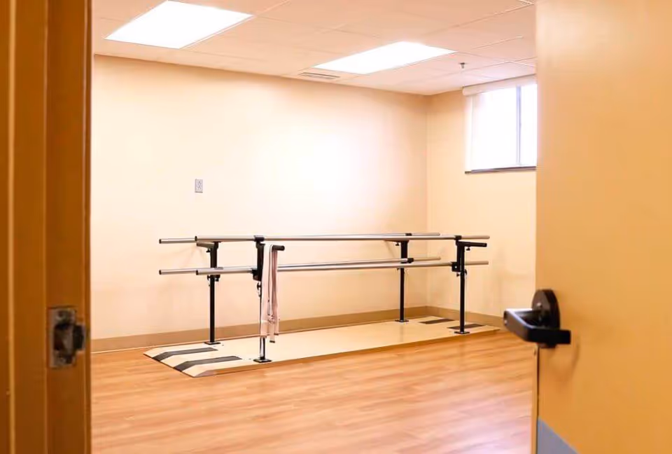 Empty rehabilitation therapy room with parallel walking bars, a towel draped over one bar, wood-look flooring and a small window.