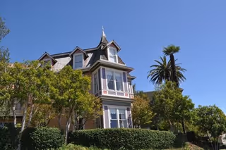 Victorian-style multi-story house with a turret surrounded by trees and palm trees under a clear blue sky.