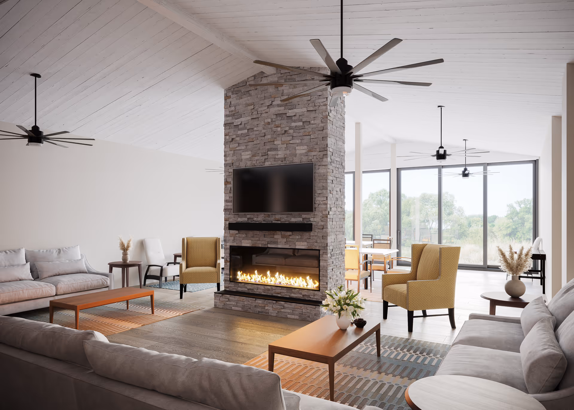 A spacious senior living common area featuring a modern stone fireplace with a flat-screen TV mounted above it. The room includes comfortable seating with gray sofas, yellow armchairs, wooden coffee tables, and side tables with decorative vases. Large windows in the background provide natural light and a view of greenery outside. Ceiling fans hang from the white wooden ceiling.