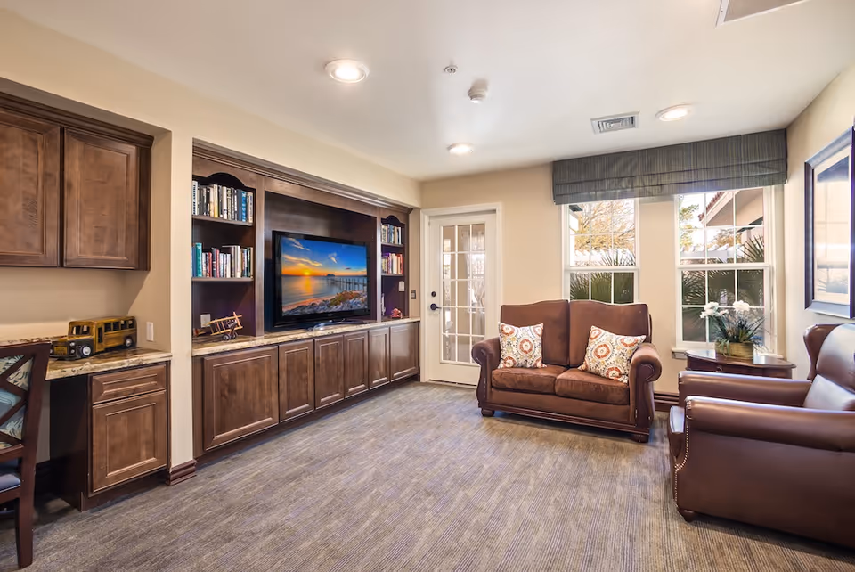 A cozy living room in a senior living facility featuring a brown loveseat with two patterned pillows, a brown leather armchair, a built-in wooden entertainment center with shelves filled with books and a flat-screen TV displaying a sunset over water. There is a small desk area with a chair and a toy vehicle on the counter. The room has beige walls, carpeted floor, recessed ceiling lights, and windows with a view of outdoor greenery.