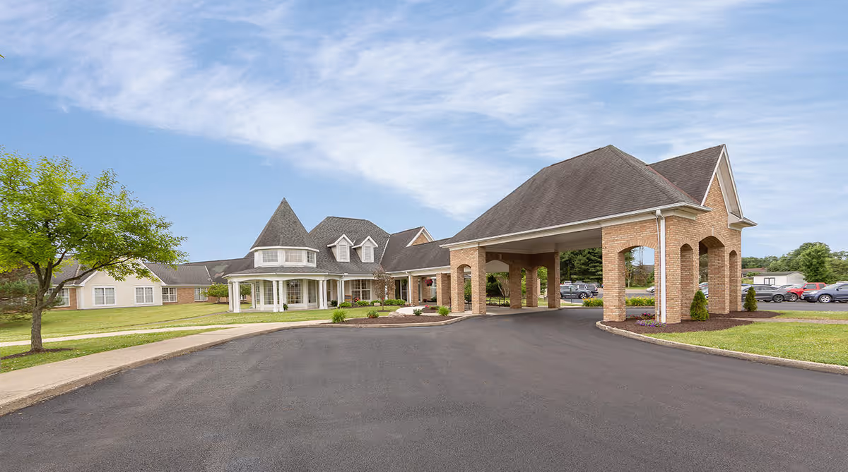 Front exterior of a senior living facility with a covered porte-cochere, turreted entrance, lawn, and parking area.