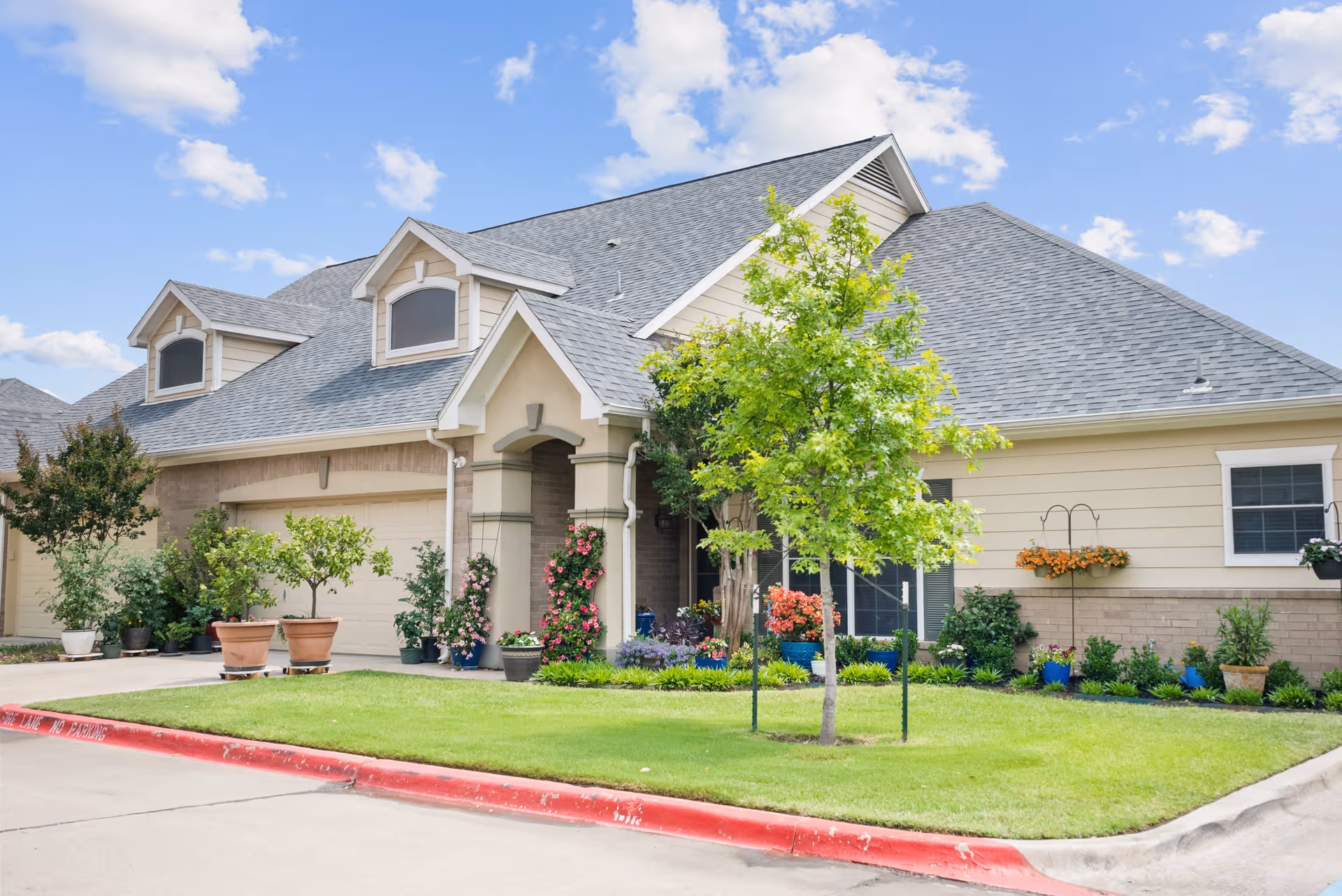 Exterior view of a single-story residential building with a gray shingled roof, beige siding, and a well-maintained front yard featuring a small tree, various potted plants, and flower beds under a blue sky with scattered clouds.