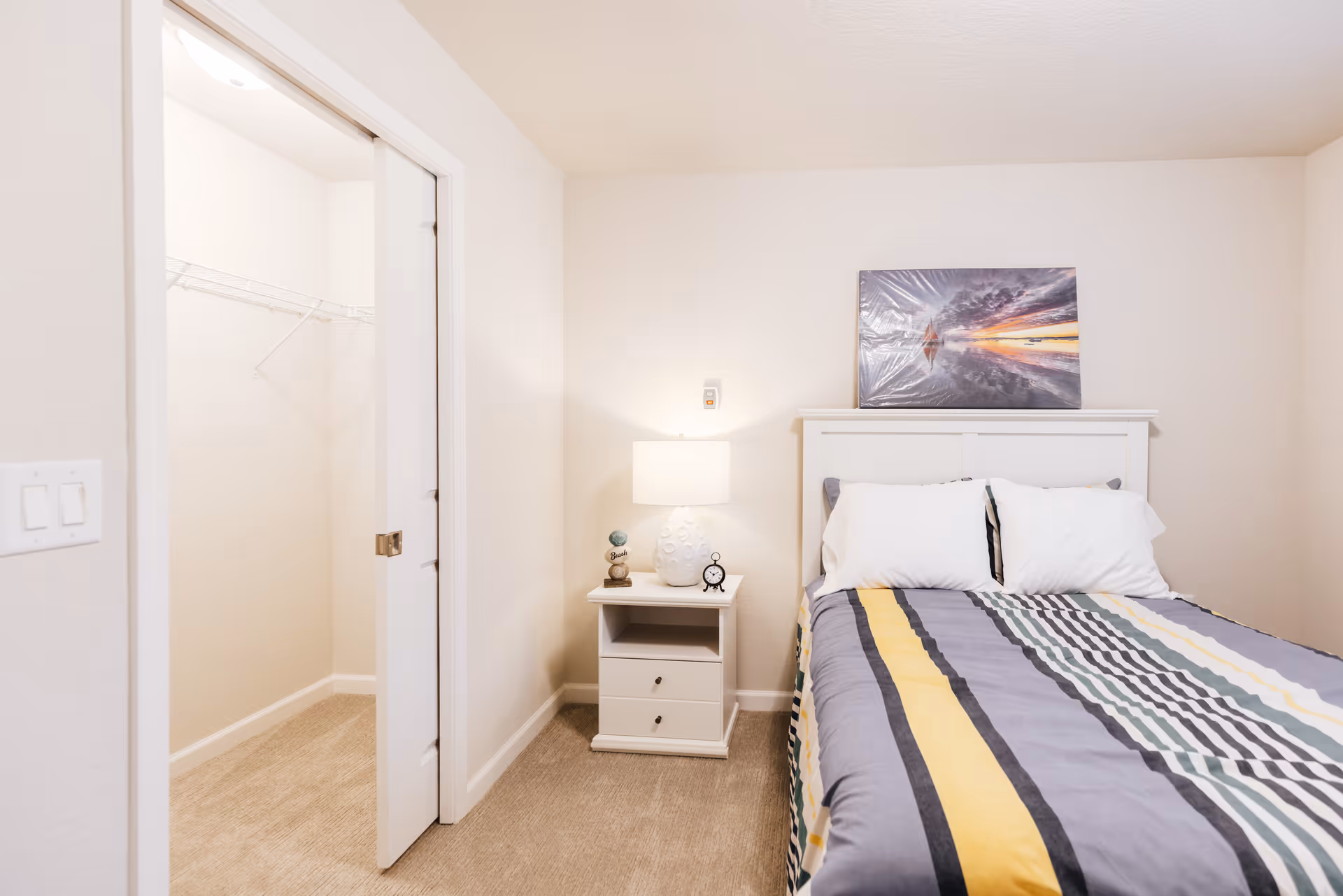 A bright bedroom with a bed featuring a striped comforter in yellow, gray, and white colors. There are two white pillows on the bed and a white headboard. A nightstand beside the bed holds a white lamp, a small clock, and decorative items. A framed picture of a sunset over water is placed above the headboard. To the left, there is an open door leading to a walk-in closet with wire shelving. The walls are light-colored and the floor is carpeted.