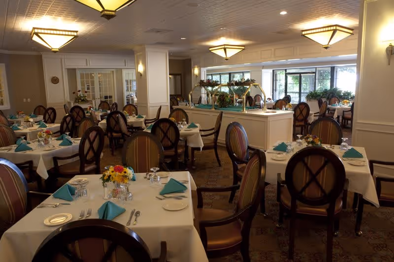 A well-lit dining room with multiple tables covered in white tablecloths, each set with turquoise napkins, plates, glasses, and silverware. The room features upholstered chairs with wooden frames, decorative light fixtures on the ceiling, and a buffet station with floral arrangements in the background.