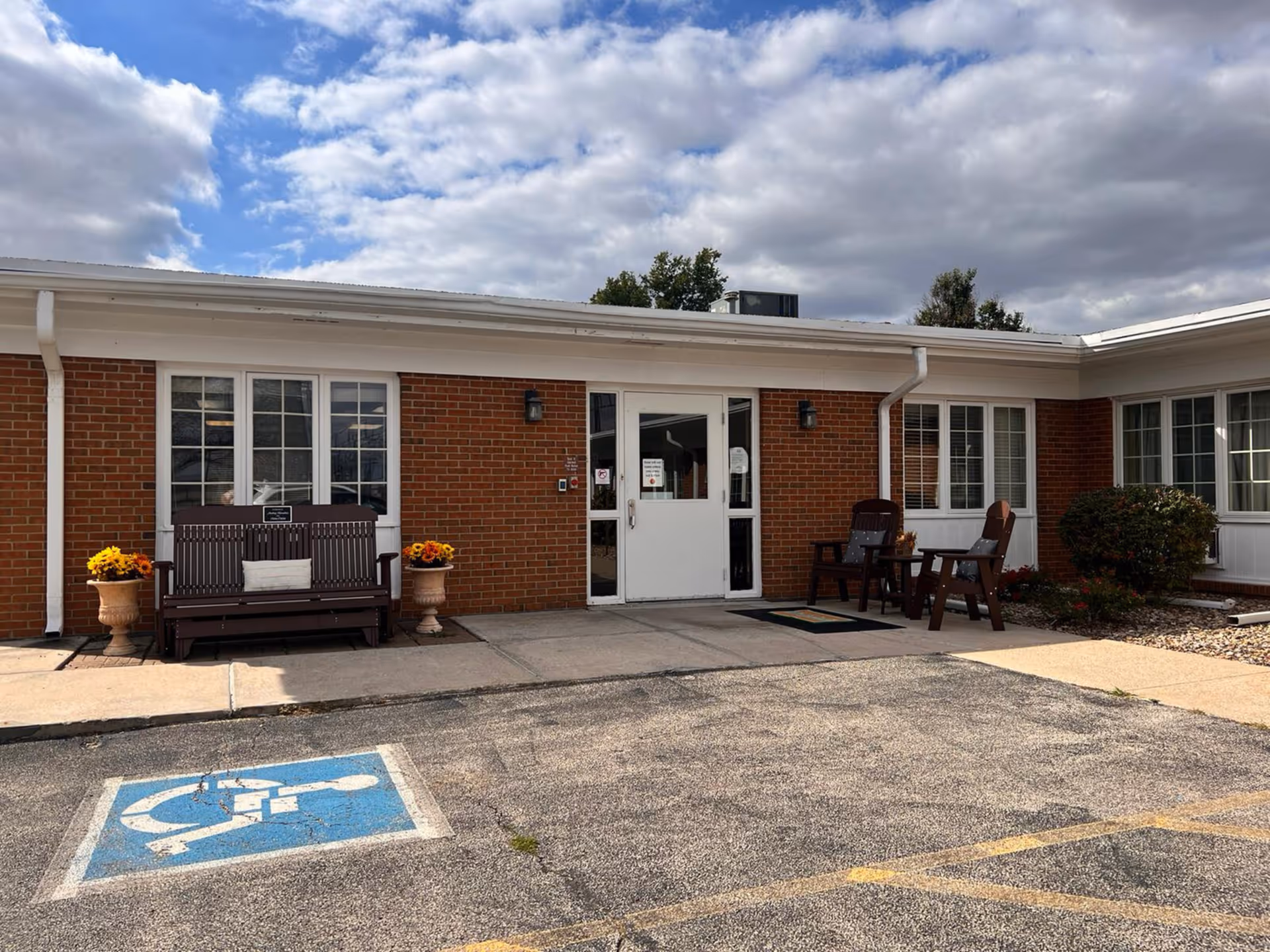 Exterior view of a single-story brick building with white trim, featuring a white double door entrance. There are two wooden benches with cushions and flower pots on either side of the entrance. A handicapped parking space is visible in the foreground. The sky is partly cloudy.