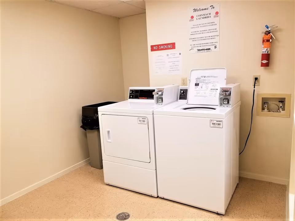 A small laundry room with a coin-operated top-loading washing machine and front-loading dryer next to a trash bin, wall-mounted fire extinguisher and signage.
