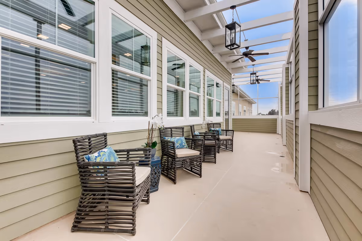 A covered outdoor patio area with four dark wicker chairs with beige cushions and blue patterned pillows lined up against a light green wall with white-framed windows. The patio has a beige floor, ceiling fans, and hanging lantern-style lights under a white pergola roof.