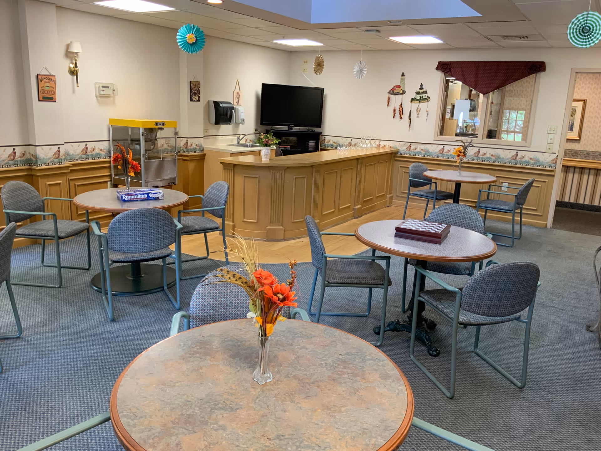 A common area in a senior living facility with several round tables and chairs arranged for socializing or games. The tables have decorative flower vases and board games like Rummikub and a chess/checkers set. There is a wooden counter with a sink and a television mounted on the wall behind it. The room is decorated with hanging paper ornaments and wall art, and has a carpeted floor with a section of wood flooring near the counter.