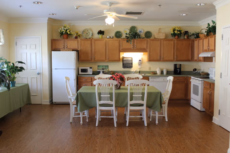 A bright kitchen and dining area with wooden cabinets, white appliances including a refrigerator, microwave, dishwasher, and stove. A green tablecloth covers a dining table surrounded by six white chairs. The kitchen counter has various decorative plants and plates displayed above the cabinets. The floor is wooden, and there is a ceiling fan with a light fixture above the table.