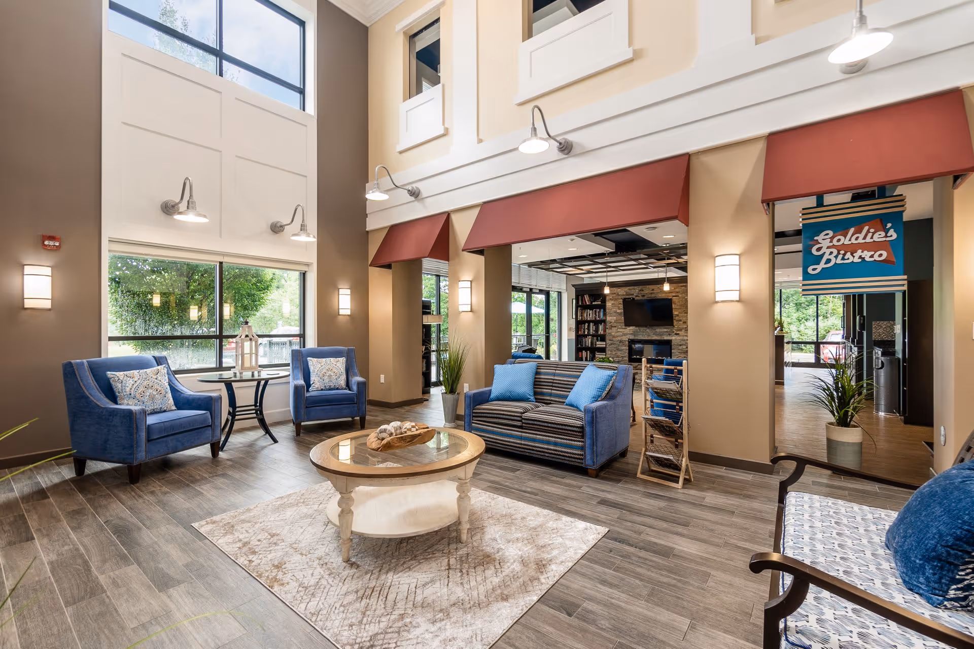 A bright and spacious senior living facility common area with high ceilings and large windows. The room features blue upholstered chairs and a loveseat with patterned cushions, a round wooden coffee table on a beige rug, and wall-mounted lights. In the background, there is a sign for 'Goldie's Bistro' above an entrance leading to another room with a stone fireplace and bookshelves.