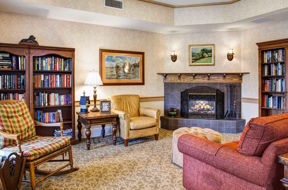 A cozy living room with a lit fireplace, two wooden bookshelves filled with books, a plaid rocking chair, a yellow armchair, a red sofa, a wooden side table with a lamp and framed photo, and two paintings on the walls.