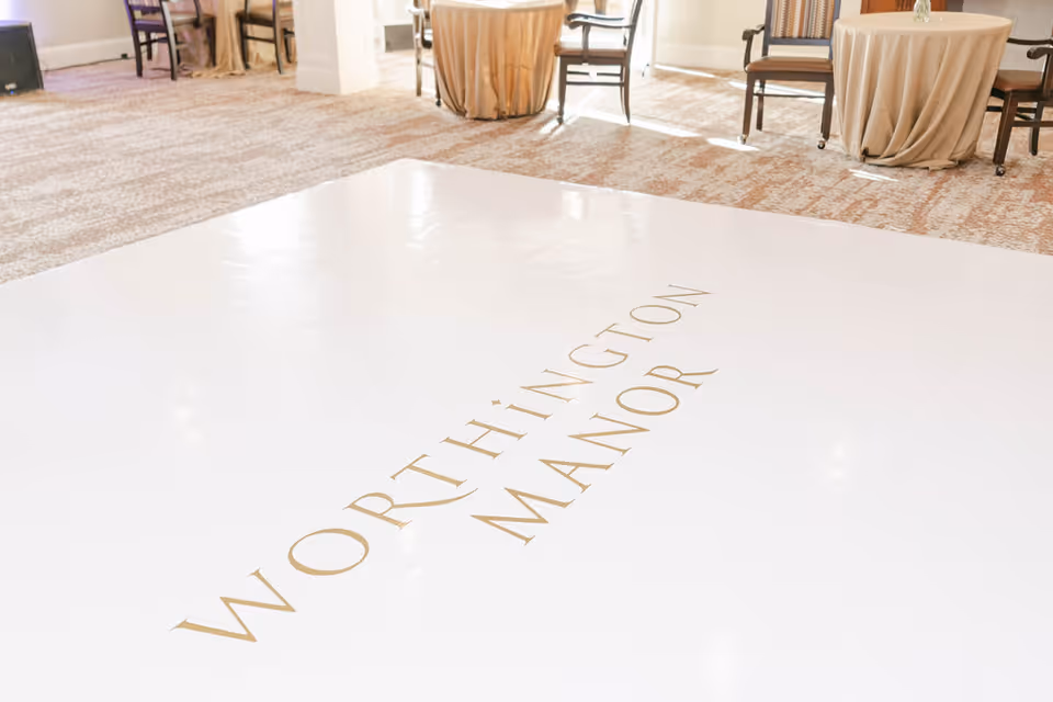 A bright room with carpeted floor and round tables covered with beige tablecloths, surrounded by chairs. The floor has a large white area with the words 'WORTHINGTON MANOR' written in gold letters.