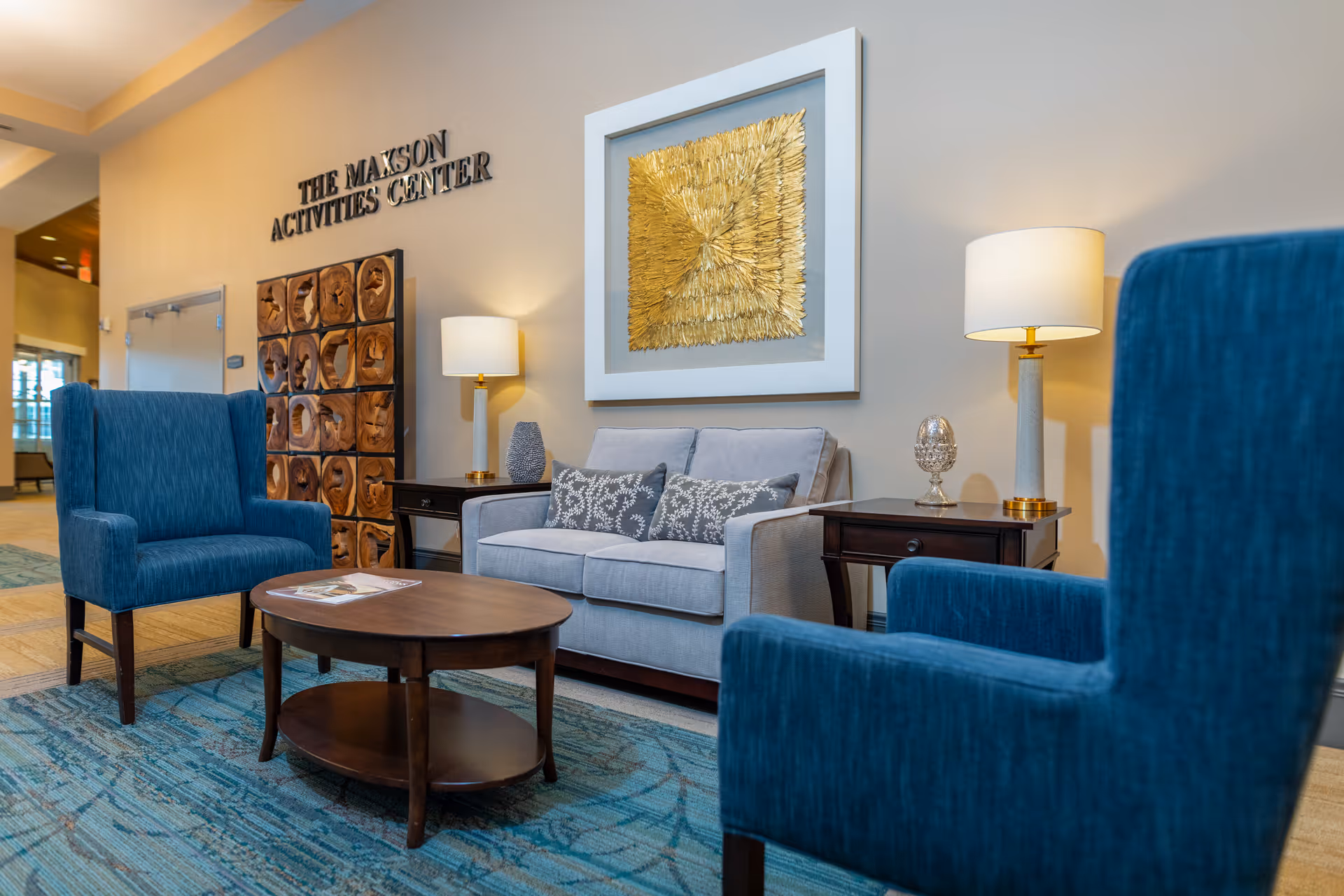 A cozy seating area in The Maxson Activities Center featuring two blue upholstered armchairs, a light gray loveseat with patterned pillows, a wooden oval coffee table, two side tables with lamps, and a decorative golden artwork on the wall.