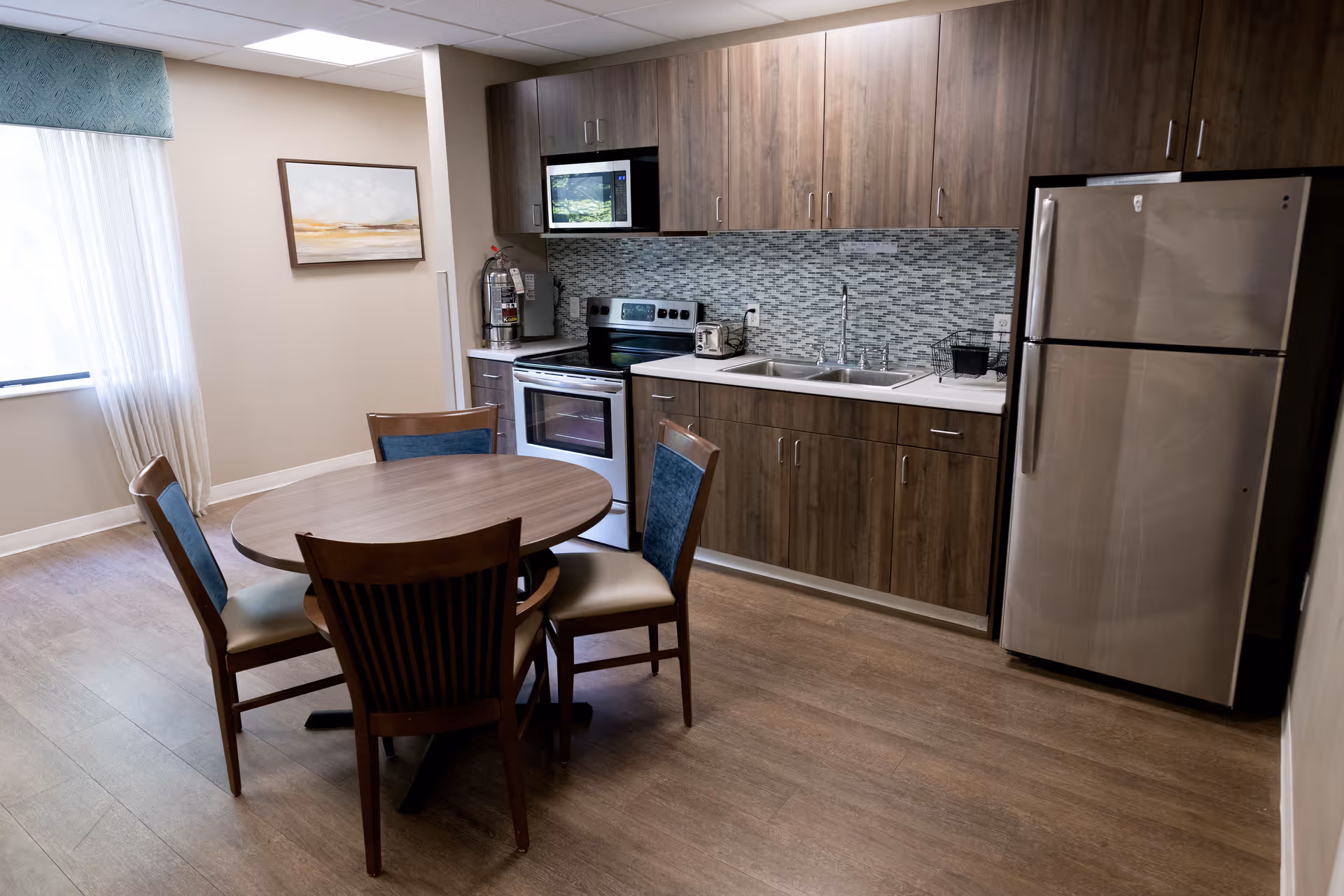 A kitchen area in a senior living facility featuring wooden cabinets, a stainless steel refrigerator, an electric stove with oven, a microwave, a double sink, and a round wooden dining table with four chairs. There is a window with sheer curtains and a framed painting on the wall.