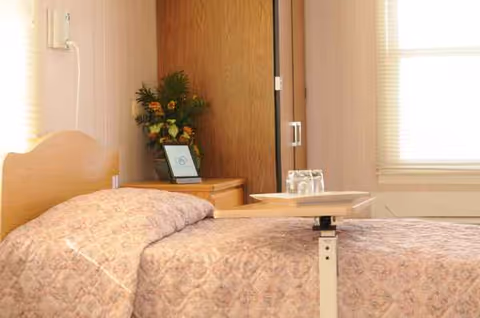 A hospital-style bedroom in a senior facility with a patterned bedspread, nightstand, and tray.