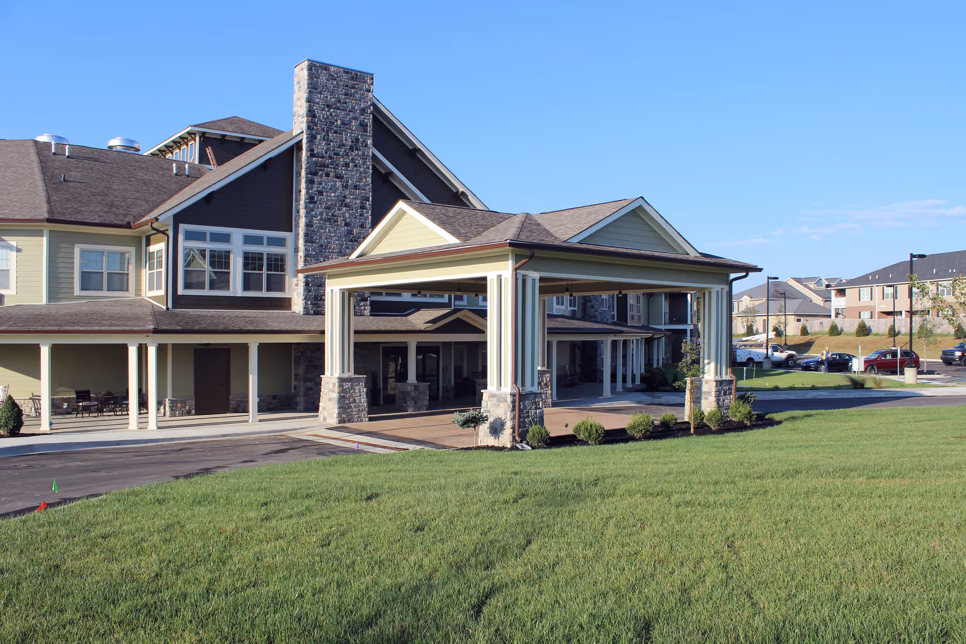 Exterior view of Dominion Senior Living of Frankfort showing a large covered entrance with stone pillars, a multi-story building with beige and dark brown siding, multiple windows, and a well-maintained green lawn in the foreground under a clear blue sky.