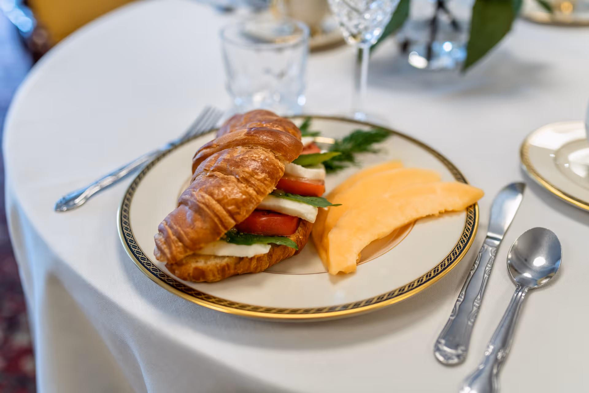A close-up of a plate on a white tablecloth with a croissant sandwich filled with tomato, mozzarella, and basil, accompanied by slices of cantaloupe melon. Silverware including a fork, knife, and spoon are placed around the plate, with a glass of water and a wine glass in the background.