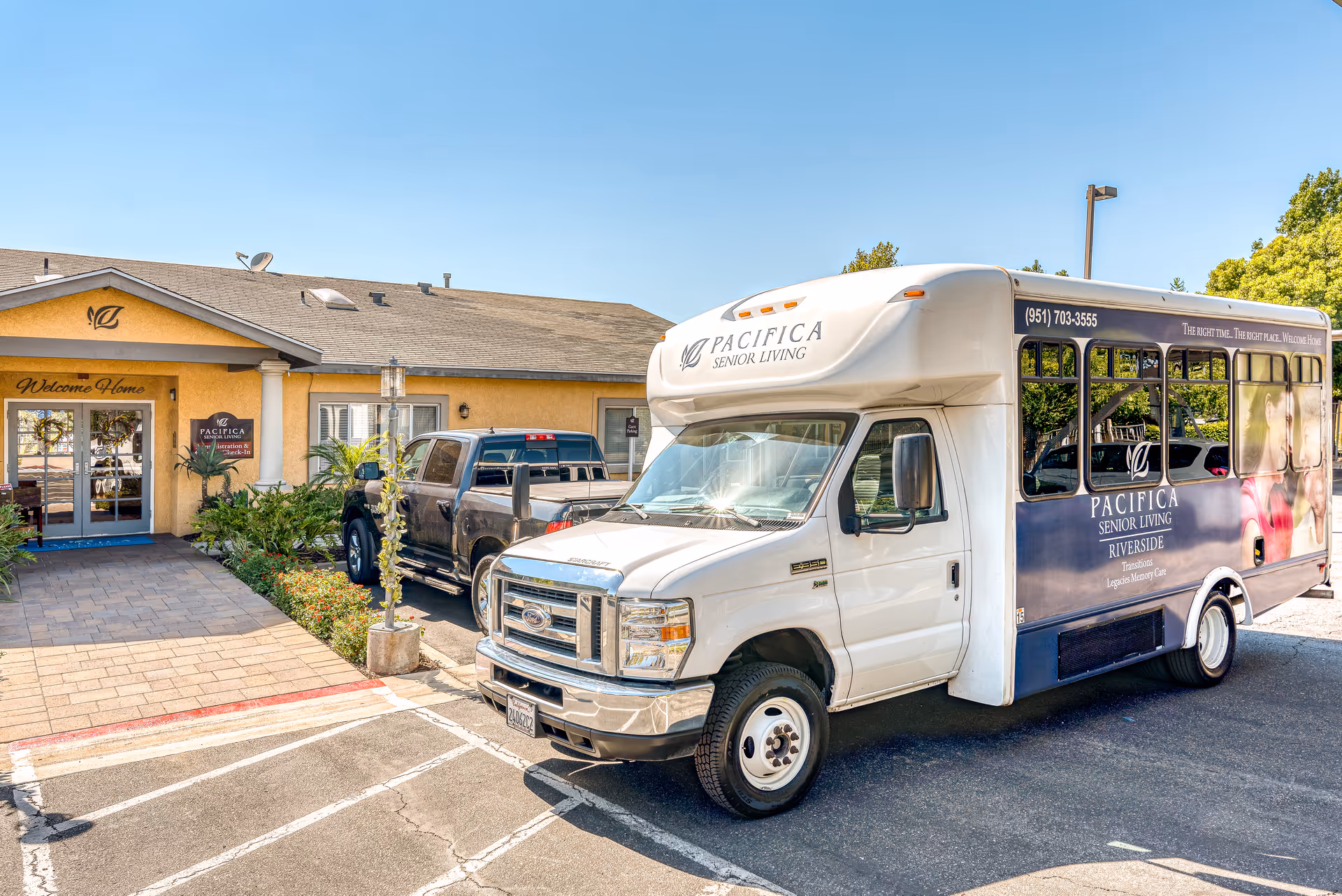 A Pacifica Senior Living shuttle van parked in front of a senior living facility entrance with landscaping and a pickup truck nearby.