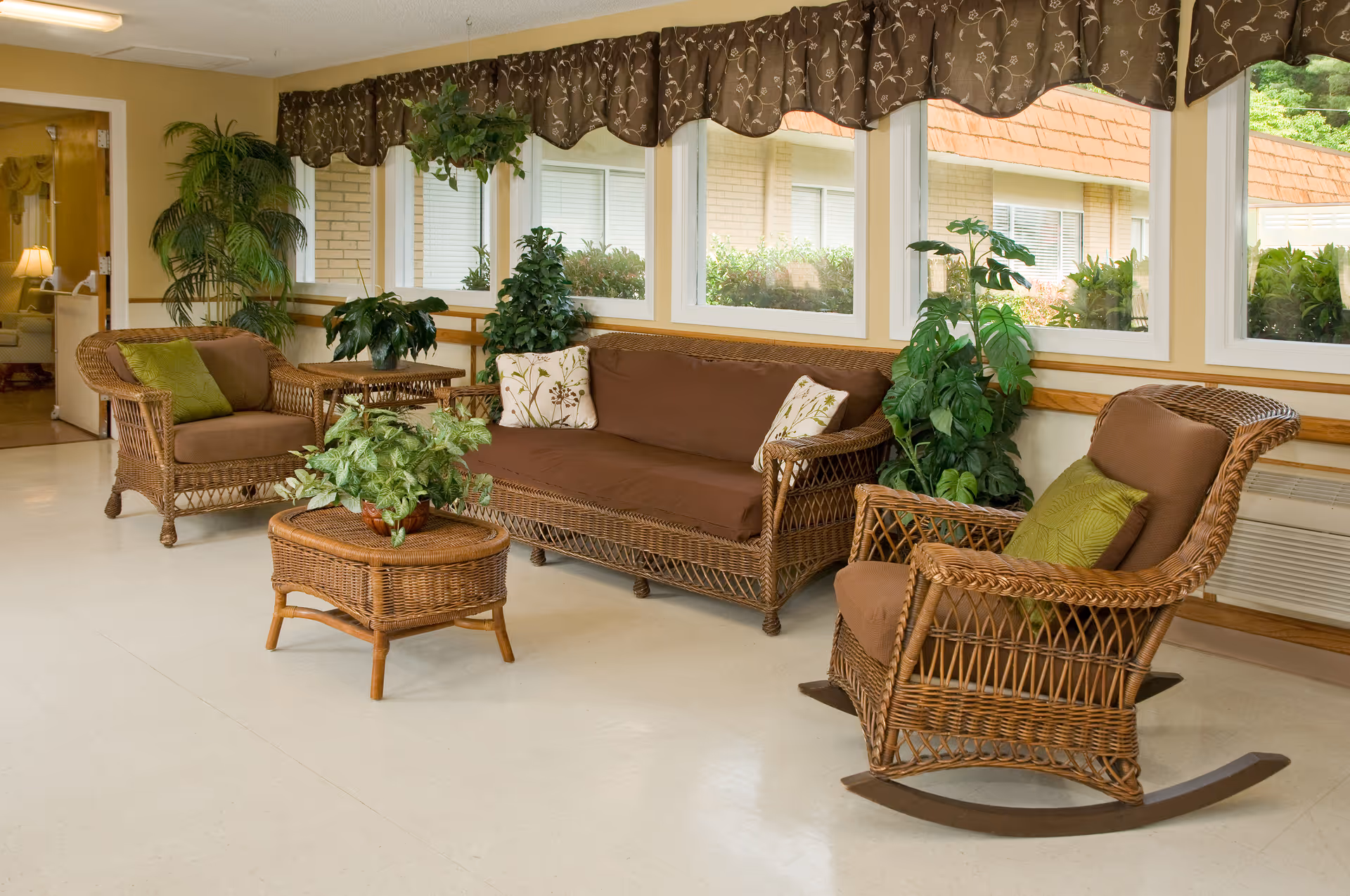 Wicker seating area with brown cushions, a rocking chair, coffee table and potted plants in a bright sunlit lounge with large windows.