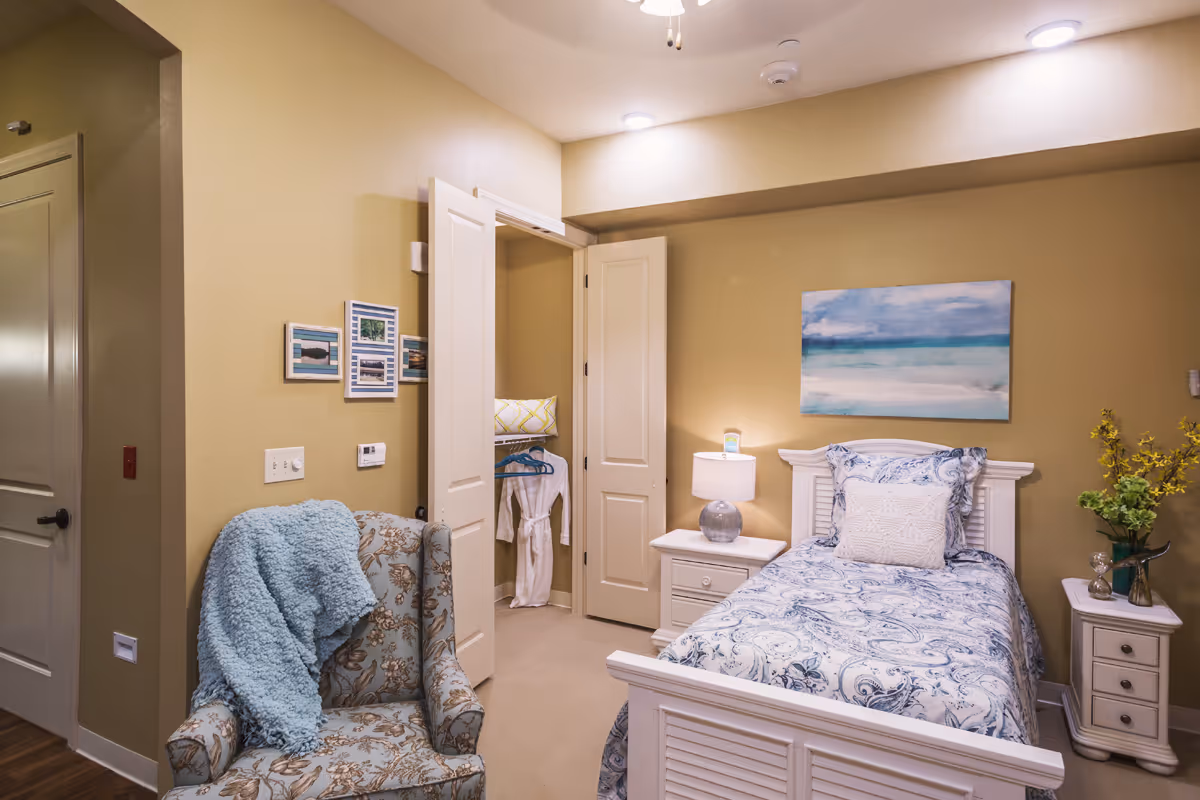 A cozy bedroom with a single bed featuring blue and white patterned bedding and a white headboard. There is a nightstand with a lamp and a small device on it next to the bed. A floral armchair with a blue throw blanket is positioned near the bed. The room has beige walls with a painting of a seascape above the bed. An open closet door reveals hanging clothes and a pillow inside. A small table with a vase of yellow flowers is also visible.