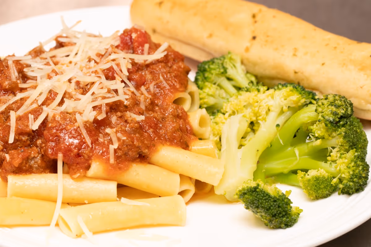A plate of food featuring pasta with meat sauce topped with shredded cheese, steamed broccoli, and a breadstick.