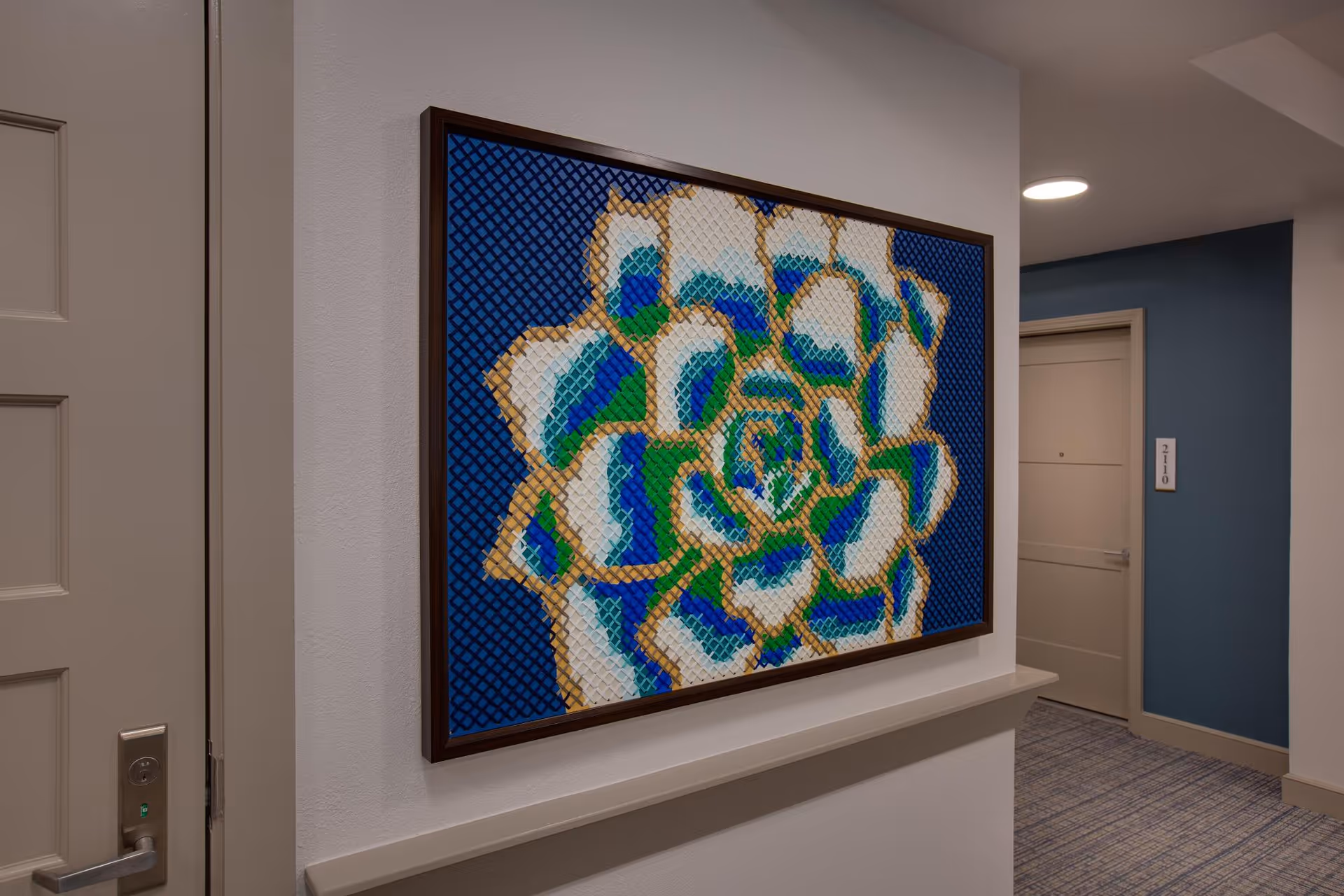 A hallway in The Hacienda at Georgetown featuring a large framed abstract artwork with blue, green, white, and gold colors on the wall. The hallway has beige doors and blue accent walls with carpeted flooring.