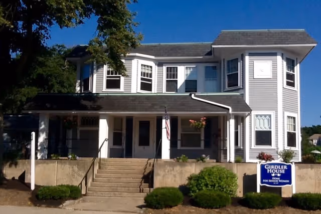 Front view of a two-story gray Victorian-style house with a covered porch, central steps, and a sign reading 'Girdler House'.