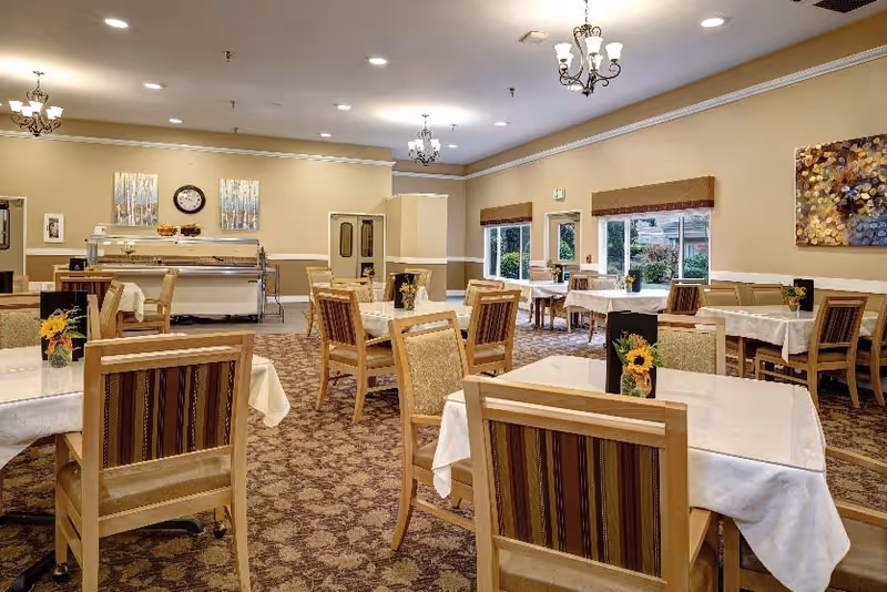 A dining room in a senior living facility with multiple tables covered in white tablecloths, each surrounded by wooden chairs with striped cushions. The room has beige walls, carpeted floors, and several windows letting in natural light. There are chandeliers hanging from the ceiling and decorative artwork on the walls.