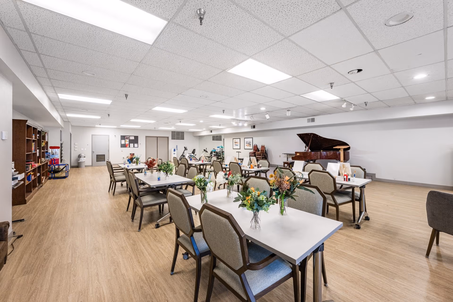 A spacious, well-lit activity or multipurpose room with several tables and chairs arranged in rows. Each table has a small vase with colorful flowers. In the background, there is a grand piano, some stacked chairs, and exercise equipment. The room has a light wood floor and a white drop ceiling with fluorescent lighting.