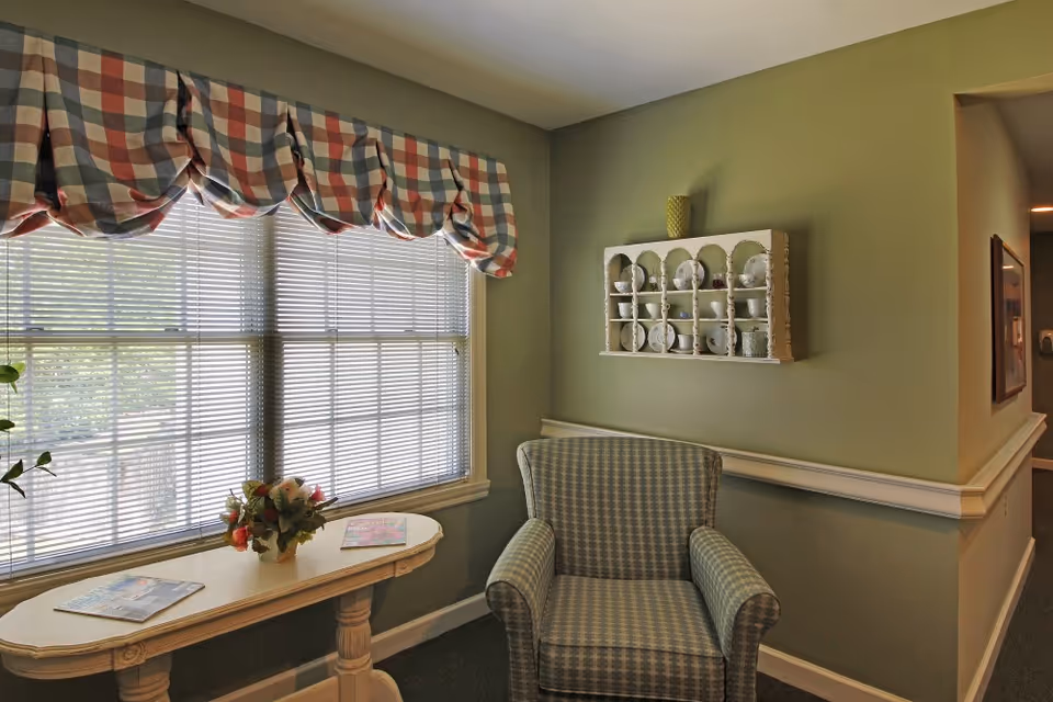 Cozy sitting area with a patterned armchair beside a table holding a floral arrangement under a large window with blinds and a checked valance, and a wall shelf displaying dishes.