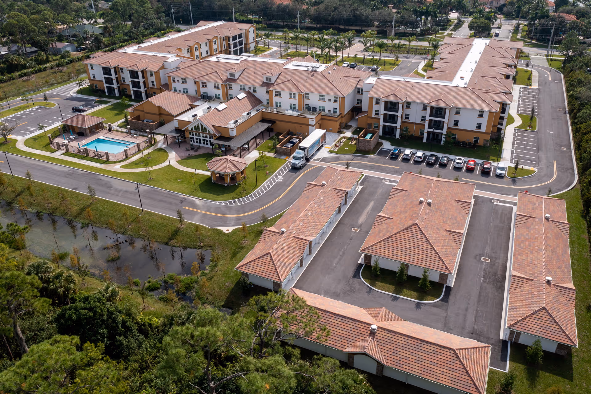 Aerial view of Palomino Gardens Retirement Community showing multiple buildings with terracotta roofs, parking lots, a swimming pool, green lawns, and surrounding trees and water features.