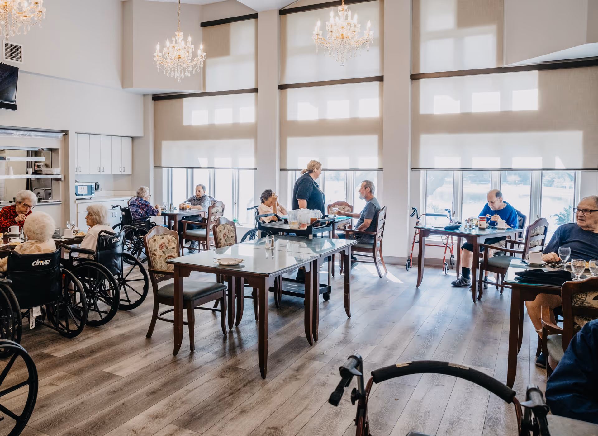 A bright dining room in an assisted living facility with several elderly residents seated at tables, some in wheelchairs, eating and conversing. A staff member is standing and interacting with one of the residents. The room has large windows with shades, wooden flooring, chandeliers, and a serving cart in the center.