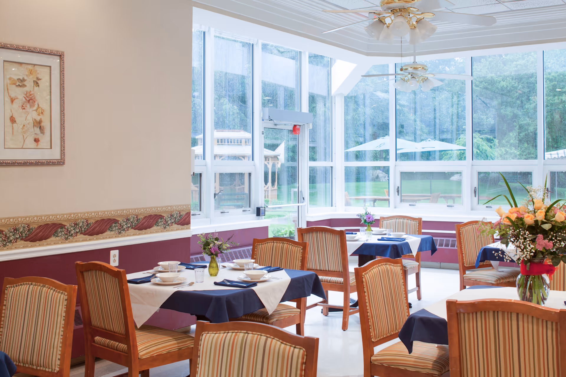 A bright dining room with several tables covered in white and navy blue tablecloths, set with plates, bowls, and napkins. The room has large windows letting in natural light and offering a view of a green outdoor area with umbrellas. There are wooden chairs with striped cushions around the tables, a ceiling fan with lights, floral decorations on the tables, and a framed floral picture on the wall.
