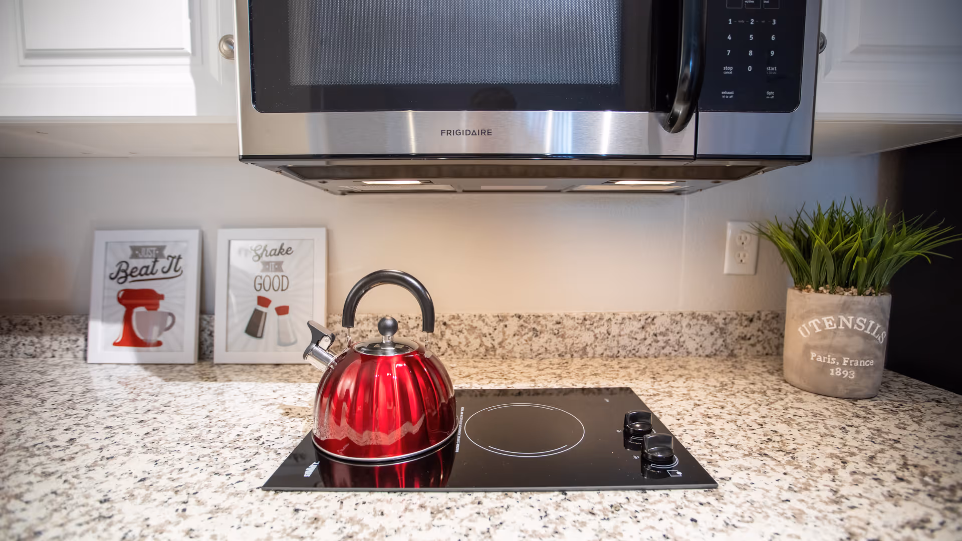 A modern kitchen countertop with a red kettle on a black electric stove. Above the stove is a stainless steel Frigidaire microwave. On the left side of the countertop are two framed kitchen-themed prints, and on the right side is a small potted plant in a container labeled 'UTENSILS Paris, France 1893'.