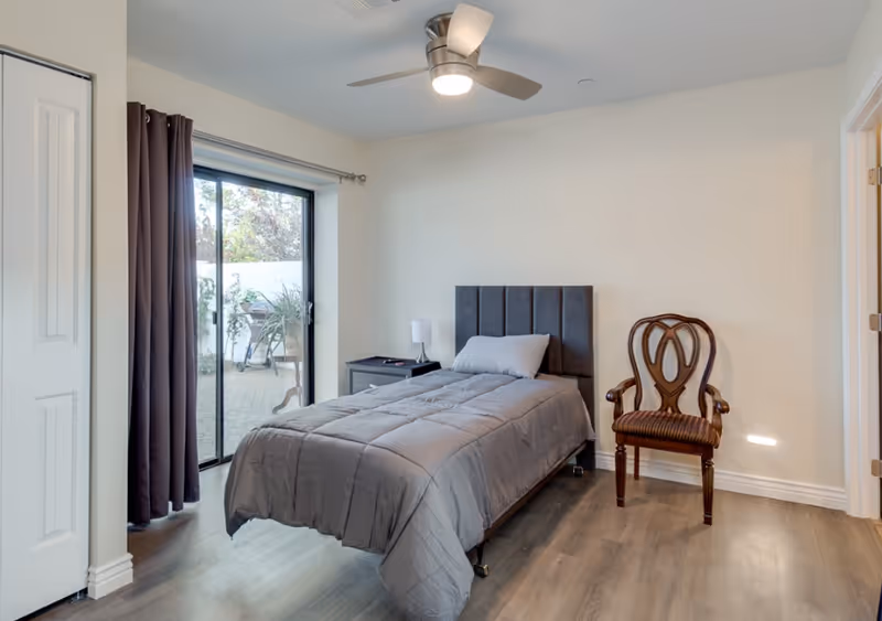 Single-occupancy bedroom with a single bed, nightstand, wooden chair, ceiling fan and sliding glass door to a patio.