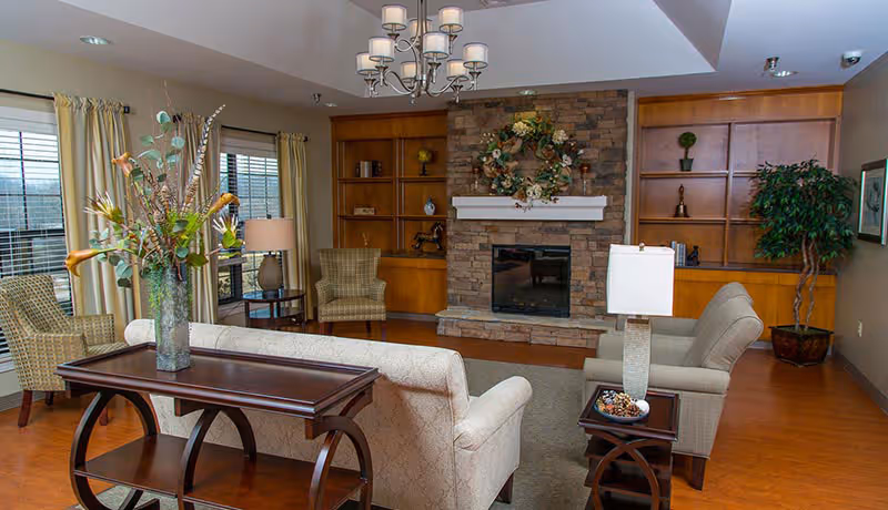 A cozy living room with a stone fireplace decorated with a floral wreath. The room features wooden built-in shelves on either side of the fireplace, two upholstered armchairs, a patterned sofa, a wooden coffee table with a vase of flowers, and a side table with a lamp. Large windows with curtains allow natural light to fill the space.
