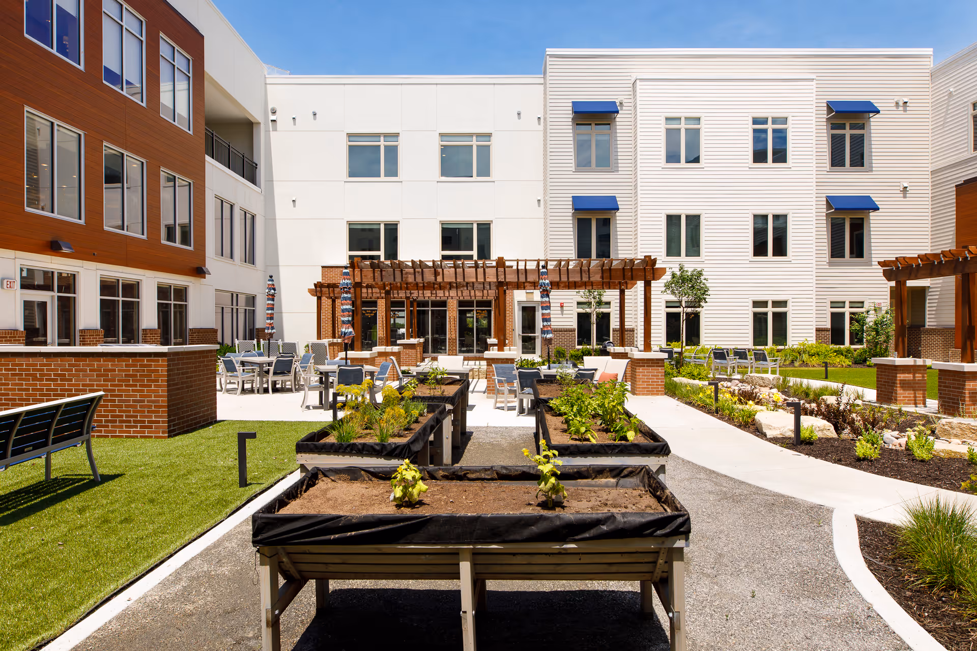 Outdoor courtyard area of a senior living facility with raised garden beds, patio seating with tables and chairs, pergolas, and a multi-story building with windows and blue awnings in the background under a clear blue sky.