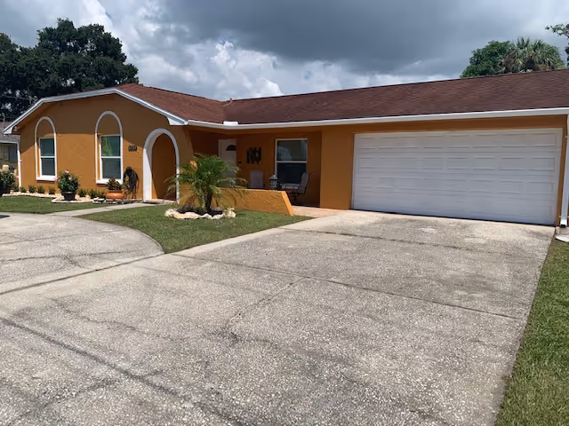 Exterior view of a single-story assisted living facility building with an orange facade, white trim, a brown shingle roof, a large white garage door, a small front porch with seating, and a driveway leading up to the garage. There is a small landscaped area with a palm plant and other greenery in front of the building under a partly cloudy sky.