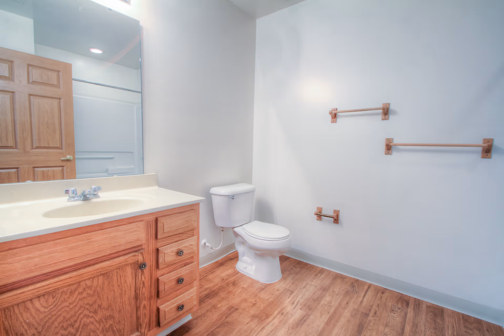 A clean bathroom with a wooden vanity featuring a sink and faucet, a large mirror above the vanity, a white toilet, and three wooden towel racks mounted on a white wall. The floor has a wood-like finish, and a wooden door is partially visible in the mirror reflection.