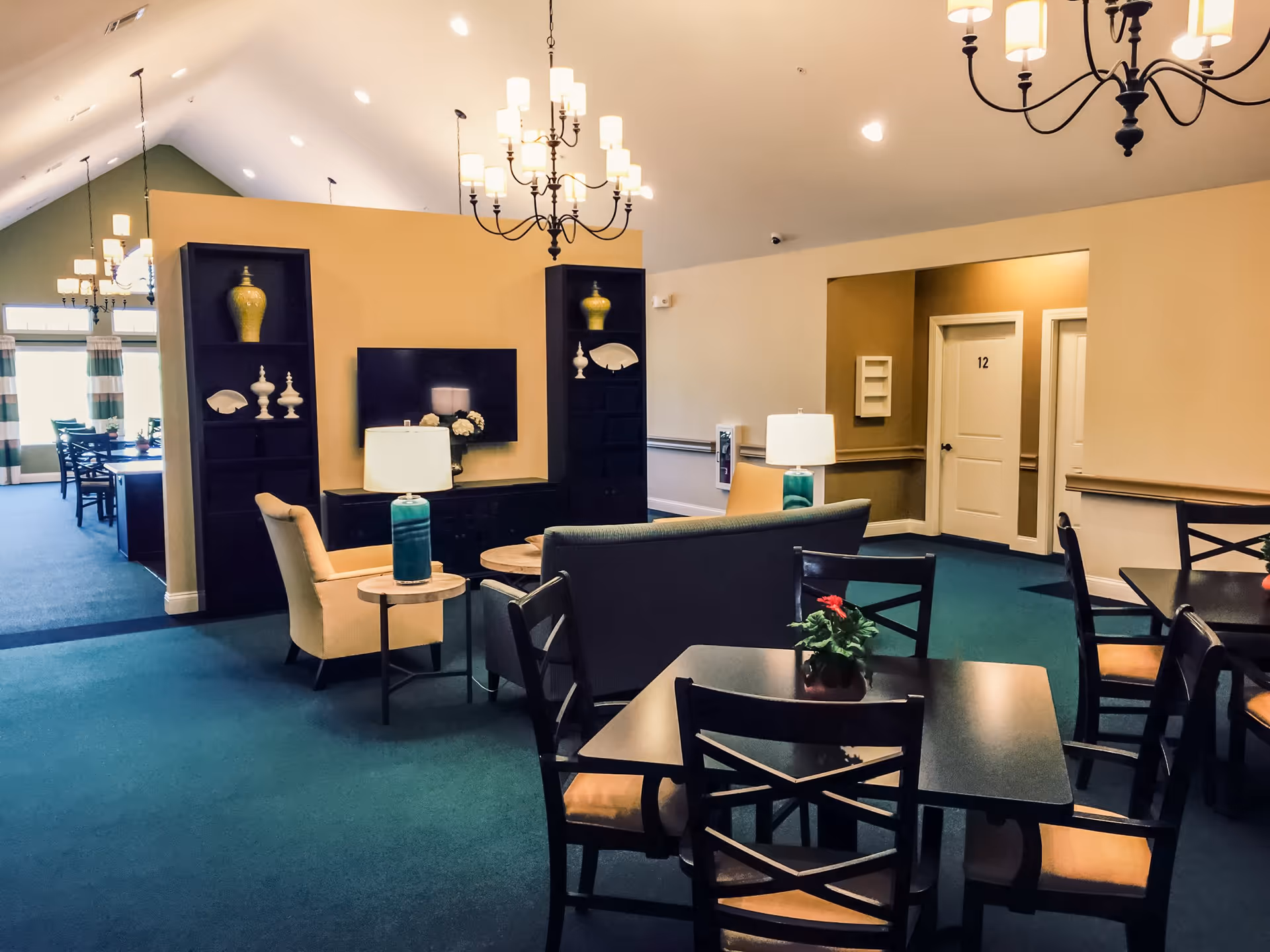 Interior view of a senior living facility common area with a seating arrangement including chairs and a sofa around a small table. There are two tall black shelves with decorative items and a flat-screen TV mounted on a beige wall. The room has green carpet flooring, chandeliers hanging from the ceiling, and a hallway with doors numbered 12.