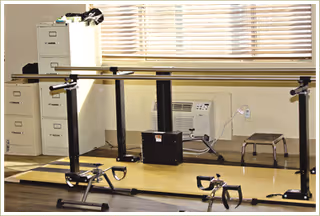 Physical therapy room with parallel walking bars, exercise equipment, mirrors, a filing cabinet, and a small stool.