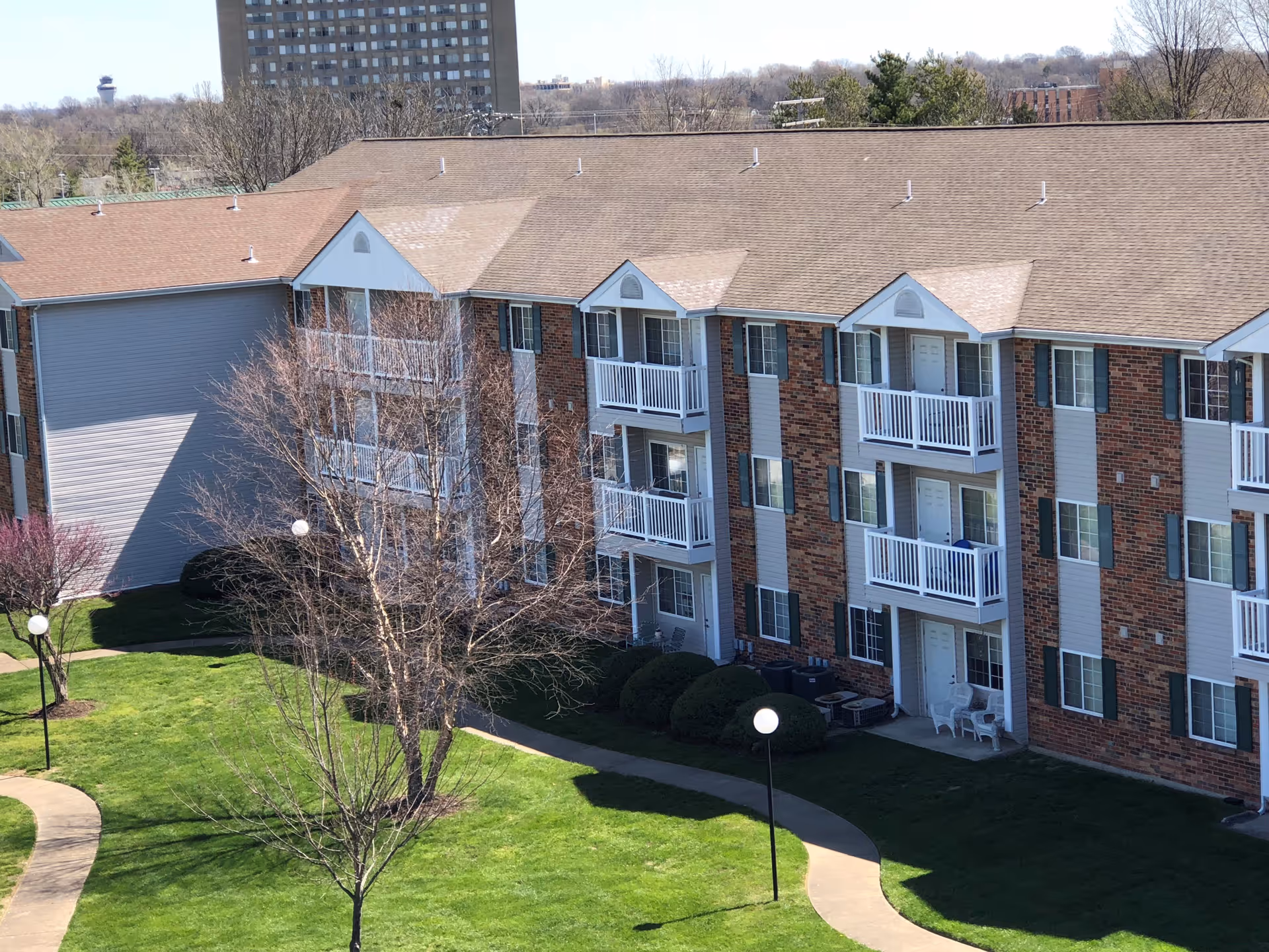 Exterior view of a three-story brick apartment building with white balconies overlooking a landscaped lawn and winding walkway.