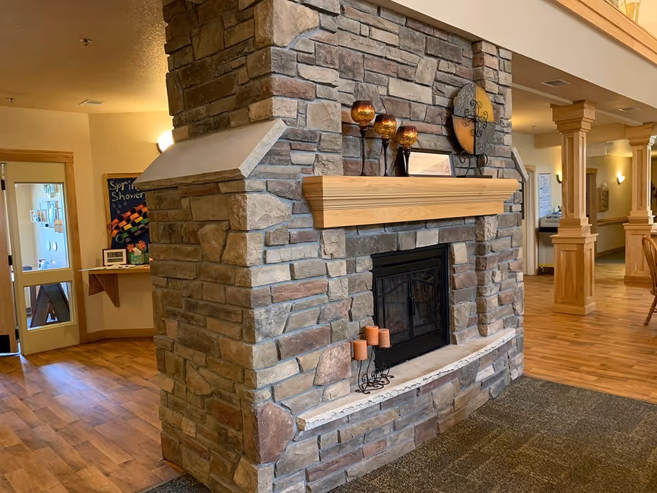 Interior view of a senior living facility featuring a large stone fireplace with a wooden mantel decorated with candles and ornamental items. The room has wooden flooring, wooden columns, and warm lighting, creating a cozy and inviting atmosphere.