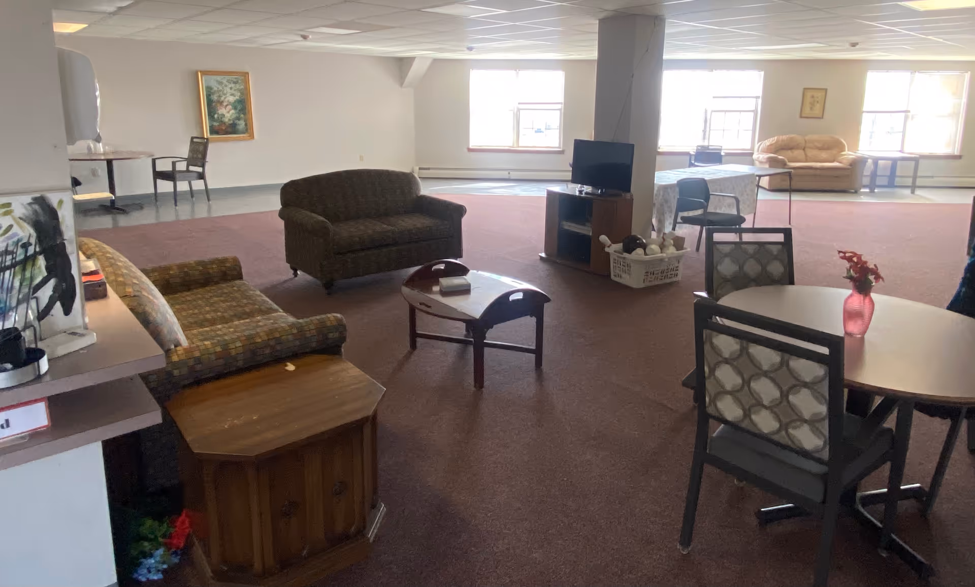Large communal lounge with sofas, armchairs, round tables and chairs, a TV on a stand, and windows letting in daylight.