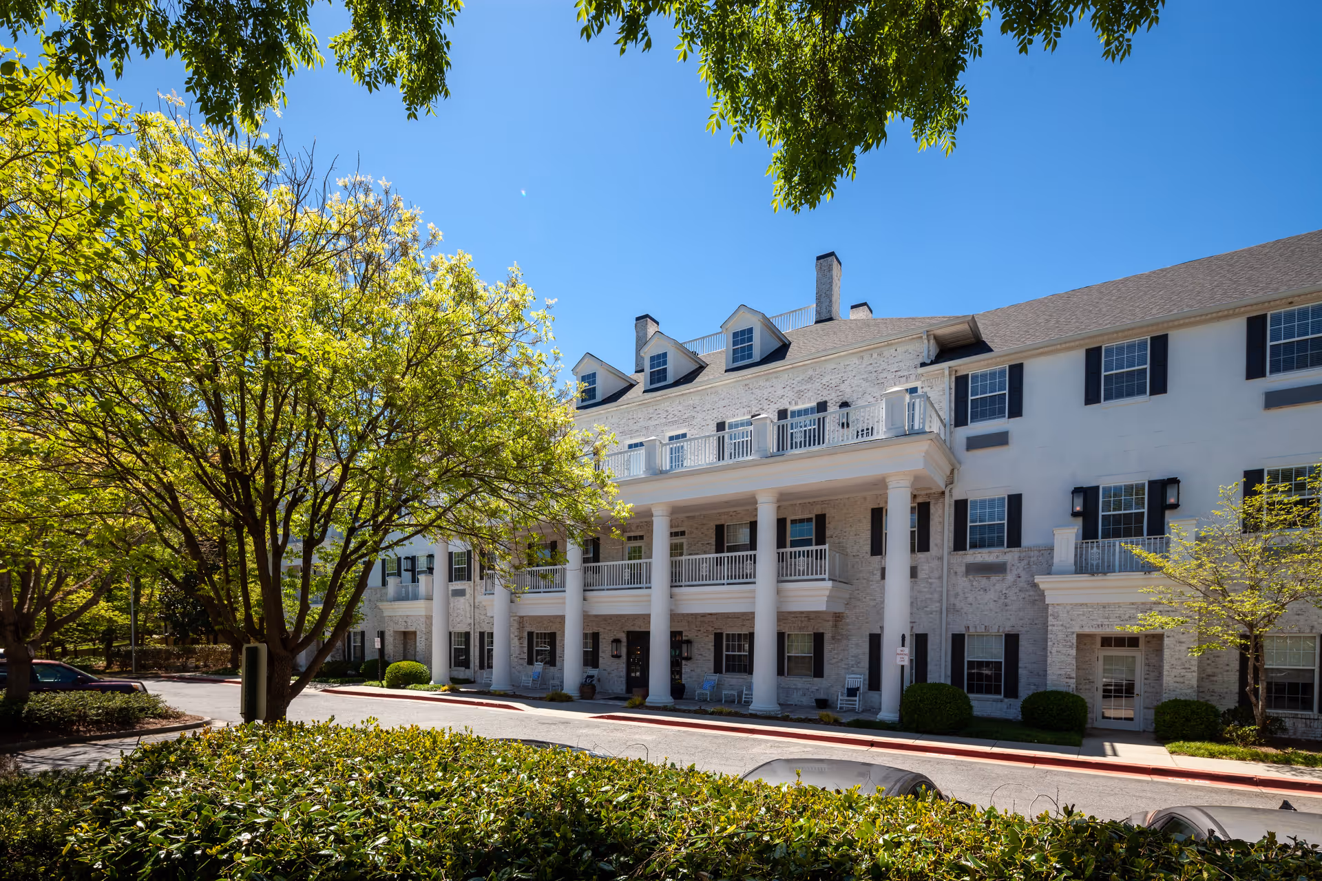 Exterior view of a three-story senior living facility building with white brick walls, black window shutters, and large white columns supporting balconies. The building is surrounded by green trees and bushes under a clear blue sky.