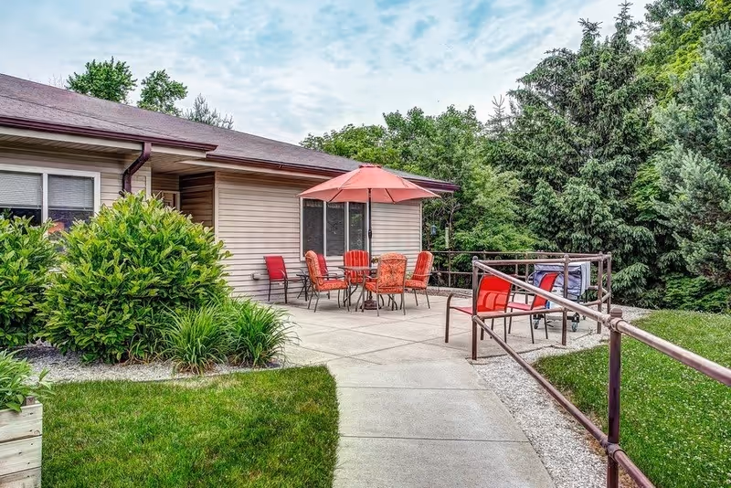 Outdoor patio area at The Waterford at West Bend with a concrete walkway, red patio chairs, a table with a red umbrella, surrounded by green bushes and trees under a partly cloudy sky.