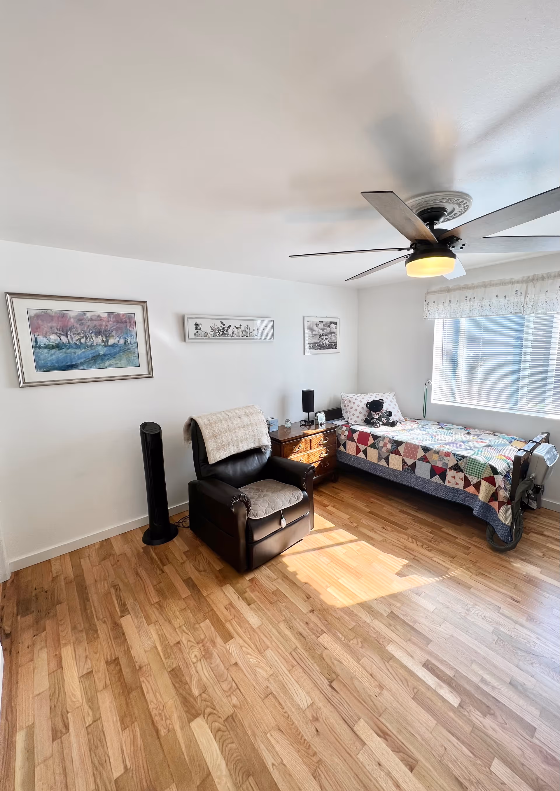 A bright bedroom with a single bed covered in a colorful patchwork quilt, a black leather recliner chair with a beige throw draped over it, a wooden nightstand with a lamp and small items, three framed pictures on the white wall, a large window with blinds and a valance, and a ceiling fan with a light fixture. The floor is wooden and sunlight streams in through the window.