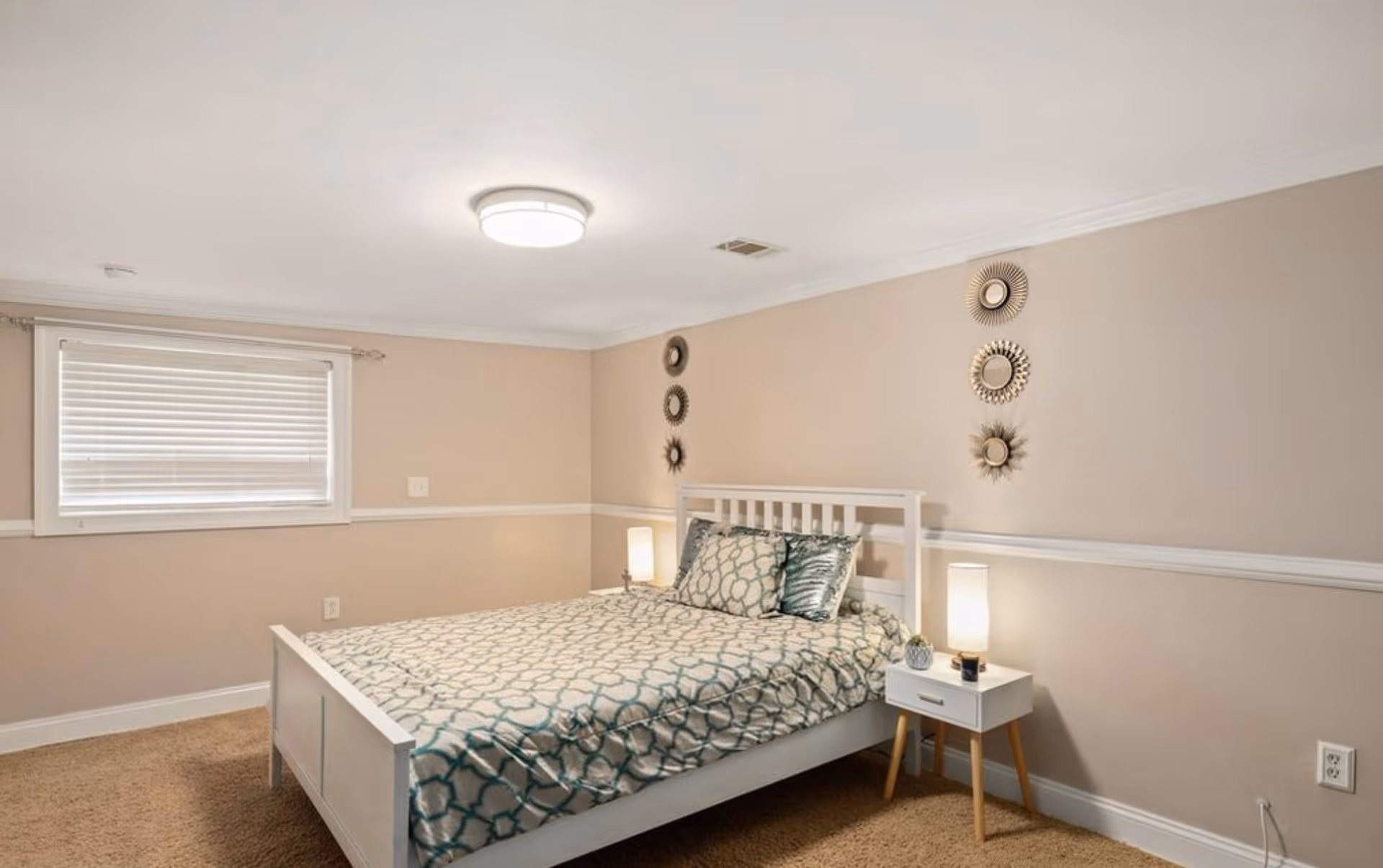 Bright bedroom with a white bed, patterned bedding, a nightstand with lamp, and decorative wall mirrors.