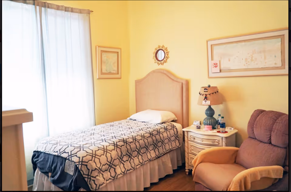 A cozy bedroom with a single bed covered in a patterned blanket, a pillow, and a beige upholstered headboard. Next to the bed is a white nightstand with a green lamp, water bottles, and small items. A comfortable armchair with yellow arm covers is positioned nearby. The room has light yellow walls with framed artwork and a small decorative mirror. A window with sheer curtains allows natural light to enter the room.