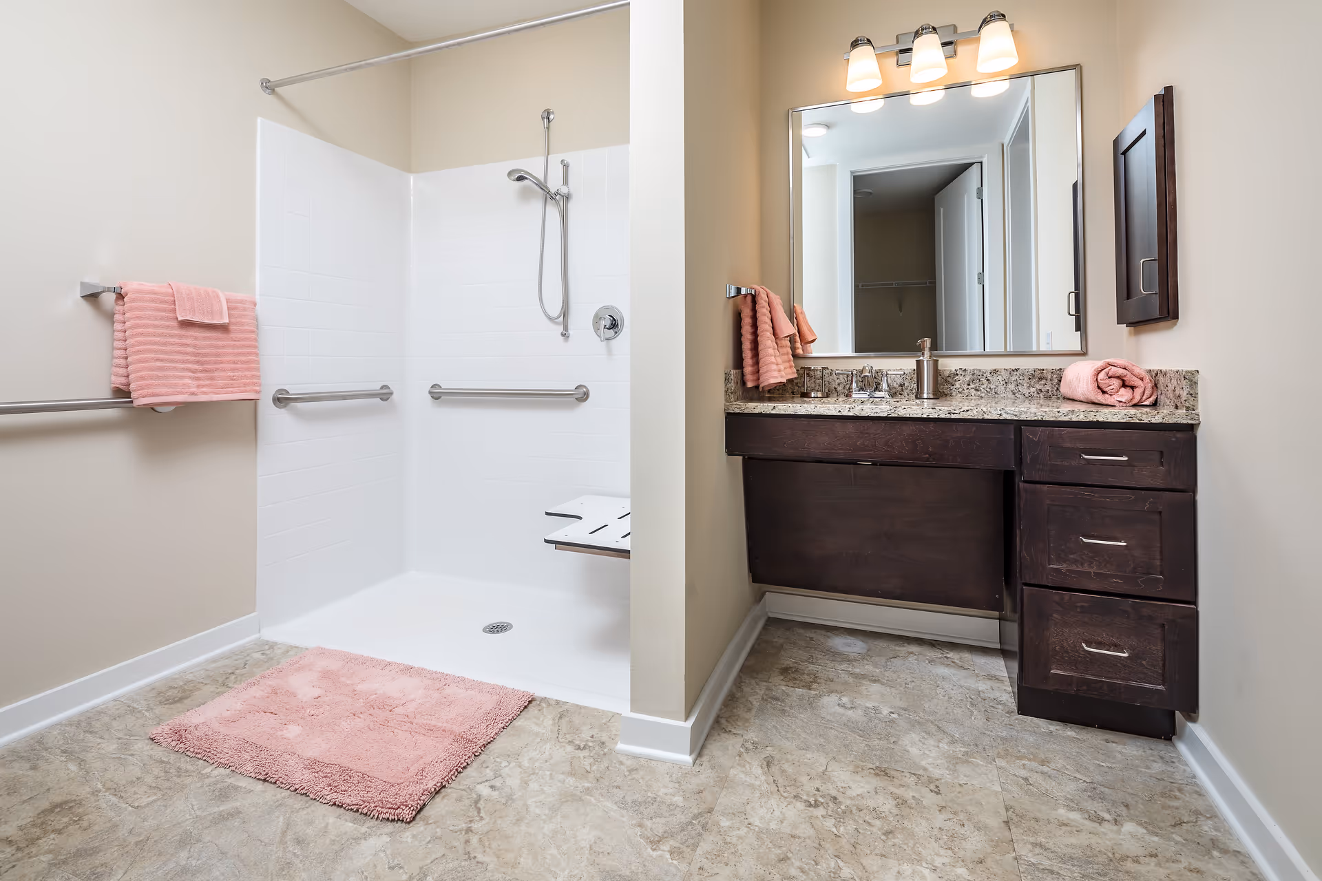 A bathroom with a walk-in shower featuring white tiles, grab bars, and a fold-down shower seat. There is a pink bath mat on the floor and pink towels hanging on a towel rack. The vanity has a granite countertop, a large mirror with three lights above it, and dark wood cabinets with drawers. A rolled pink towel is placed on the countertop.